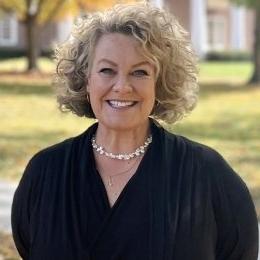 Lady with curly hair with black top and pearls standing on campus in the fall